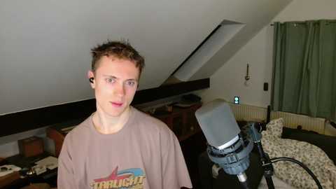 Video of a young, fair-skinned, curly-haired man wearing a beige T-shirt with a graphic, standing in a cluttered attic room with a microphone and recording equipment.
