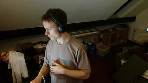 Video of a young man with short curly hair, wearing headphones, a gray T-shirt, and standing in a cluttered, dimly lit attic room with cardboard boxes and a radiator.