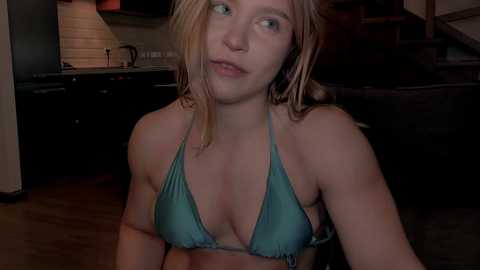 Video of a blonde woman in a teal bikini, seated indoors, looking contemplative, with a wooden floor and modern kitchen in the background.