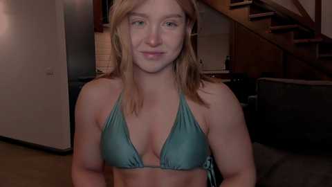 Video of a smiling young woman with fair skin and shoulder-length blonde hair, wearing a teal bikini top. She's in a dimly lit kitchen with wooden cabinets and a staircase in the background.