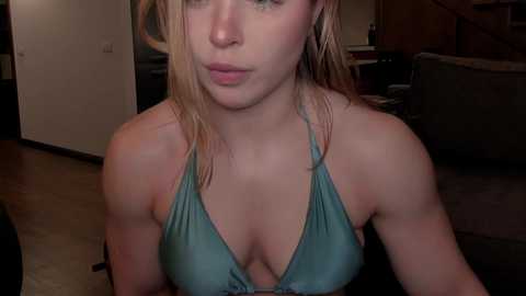Video of a young Caucasian woman with fair skin, blonde hair, and full lips, wearing a teal halter bikini top. She's indoors, on a couch, with a dimly lit background.