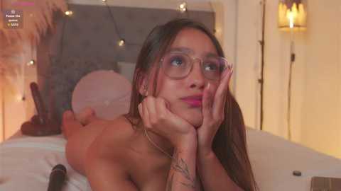 Video of a nude woman with long brown hair, glasses, and tattoos, lying on her stomach on a bed, looking thoughtful, surrounded by warm lighting and string lights.