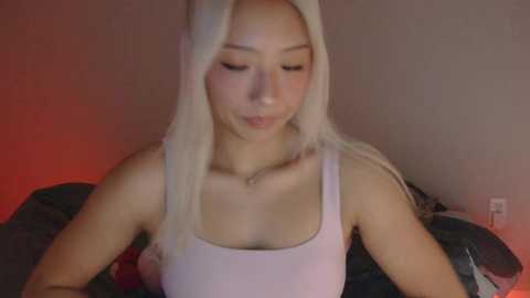 Media: A video of a young Asian woman with long, platinum blonde hair, wearing a light pink tank top, sitting on a bed with red lighting.