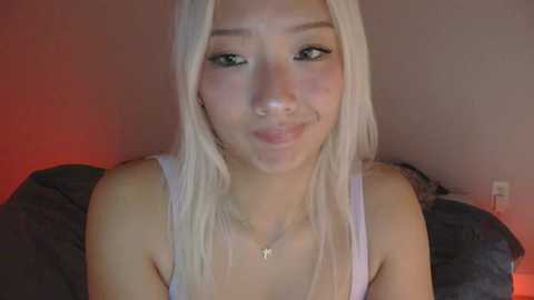Media: Video of a young Asian woman with long platinum blonde hair, wearing a white tank top, making a pouty face. She has light skin and is smiling slightly. The background is dimly lit with red and orange hues.