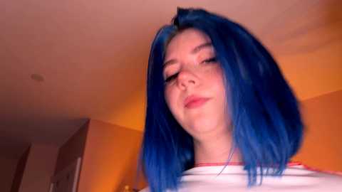 Video of a young woman with vibrant blue hair, light skin, and pink lips, wearing a white shirt. She has a serene expression. The background features warm lighting and a cozy room.