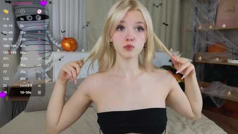 Video of a young, slender, blonde woman with fair skin, wearing a strapless black top, holding her hair. Background features Halloween decor, including a pumpkin, ghost, and spider webs.