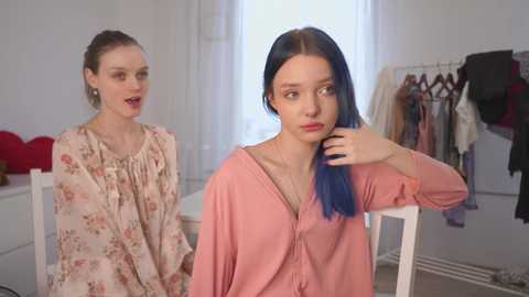 Video of two young women in a minimalist room. One with light skin and brown hair, wearing a floral blouse. Another with blue hair, light skin, and a pink blouse, resting her head on her hand.