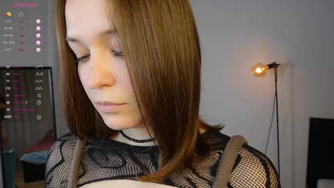 Media: Video of a young Caucasian woman with shoulder-length brown hair, wearing a black lace top, standing indoors. Background includes a monitor displaying app icons, a lamp, and blurred room details.