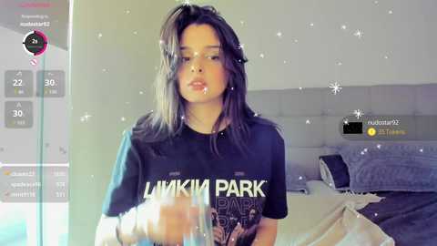 Media: A video of a young woman with long, dark hair, wearing a black \"LIVIN' PARK\" t-shirt, lying on a bed with gray pillows, in a modern bedroom with a minimalist decor.