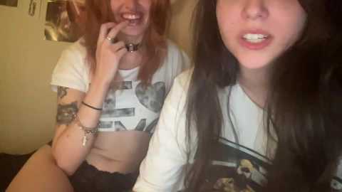 Video of two young women with fair skin, one with a white crop top and tattoos, the other in a black floral shirt, smiling at the camera.