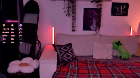 Video of a dimly lit, cozy bedroom with a white couch, green plush toy, red plaid blanket, black geometric pillow, hanging plants, and digital clock on a stand.