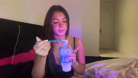 Video of a young woman with long brown hair, wearing a black bra, using a blue vibrator on herself in a dimly-lit bedroom.