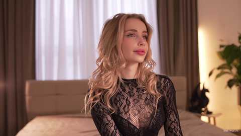 Video of a fair-skinned, wavy-haired woman with pink lips, wearing a sheer black lace top, standing in a warmly lit bedroom with beige curtains, a bed, and potted plants.