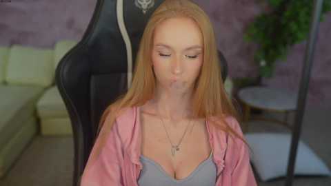 Media: Video of a fair-skinned, blonde-haired woman in a gray bra under a pink jacket, blowing bubbles, sitting in a black gaming chair. Background shows a beige couch and purple wall.