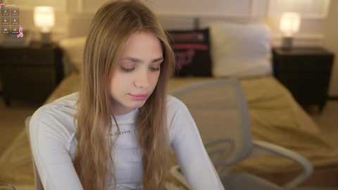 Video of a young, fair-skinned girl with long, straight blonde hair, wearing a white long-sleeved shirt, sitting on a beige bed with a cream-colored lampshade.