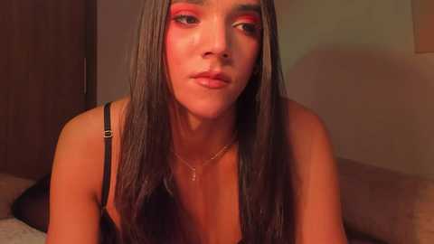 Video of a young woman with long, straight dark hair and fair skin, wearing a black bra and bold red eyeshadow. She has a neutral expression, set against a warm-toned, dimly lit bedroom background.