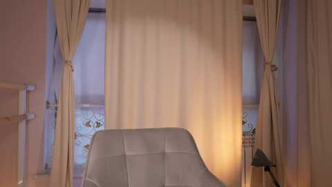 Video of a cozy, minimalist bedroom with a cream-colored upholstered chair, beige curtains, and a small, black table lamp on the right.