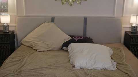 A video of a minimalist bedroom with a beige upholstered headboard, two white pillows, a black pillow with a pink flower design, and dark wooden nightstands with white lamps.