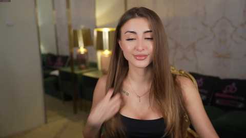 Video of a young Caucasian woman with long, straight brown hair, wearing a black strapless top, smiling in a warmly lit, ornate room with a green couch, pink pillows, and a gold lamp.