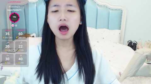 Video of a young Asian woman with long black hair, wearing a white robe, lying on a bed in a softly lit room, eyes closed, mouth open.