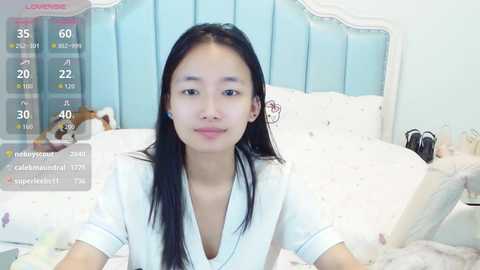 Video of an East Asian woman with long black hair, wearing a white blouse, lying on a bed with a light blue headboard and pastel-colored bedding. The room has a soft, feminine atmosphere.