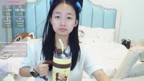 Video of an Asian woman with long black hair in a white top, holding a glass of chocolate milk with a straw, sitting on a white bed in a pastel room.