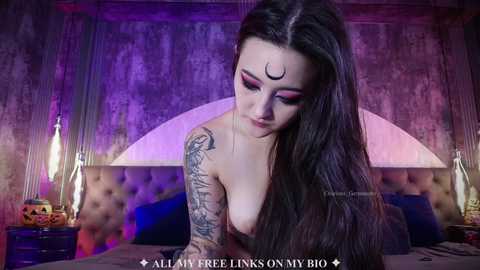 A video of a topless, tattooed woman with long black hair and a crescent moon mark on her forehead, lying on a bed with a tufted headboard, purple lighting, and a watermark reading \"ALLAH FREE UNLOCKED BY @GABRIELLA.G.\