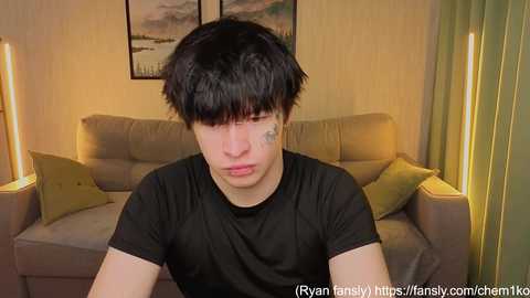Video of a young man with messy black hair, wearing a black T-shirt, sitting on a beige couch in a dimly lit living room.
