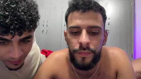 Video of two shirtless men, one with curly hair, the other with a beard, in a locker room with metal lockers and purple lighting.
