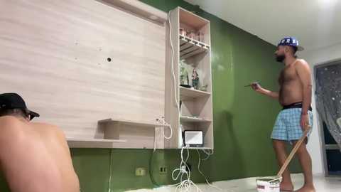 Video of two shirtless men in a minimalist green room with a white wall and wooden shelves. One man, with a black cap, kneels on the floor, while the other, wearing a blue and white cap, stands holding a broom.