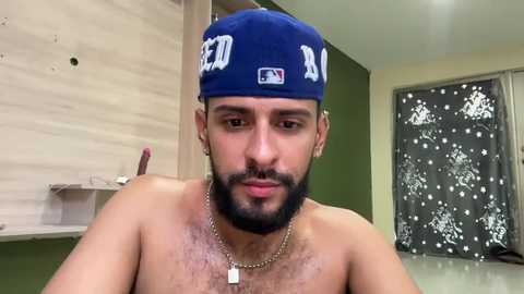 Video of a muscular, bearded man with a blue baseball cap and a necklace, in a dimly-lit room with beige walls, a shelf, and a window with floral curtains.
