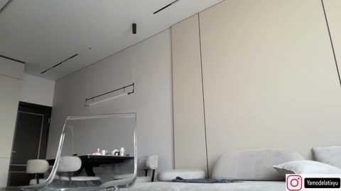 Media: Video of a minimalist bedroom with beige walls, a large mirror, and two white chairs. The room features a modern design with a large bed, a glass desk, and a neutral color palette.