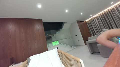 Media: Video of a modern, minimalist bedroom with a beige sofa, wooden wall, white bed with white pillows, and beige curtains; a person's hand holding a smartphone is visible.