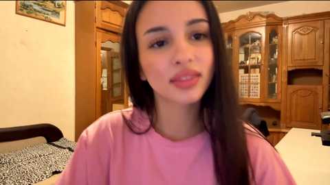 A video of a young woman with long, dark hair and fair skin, wearing a pink t-shirt, sitting in a wooden-paneled room with a bookshelf and framed painting in the background.