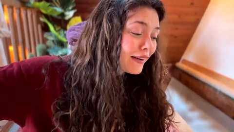 Video of a young woman with long, curly dark hair, smiling, wearing a burgundy sweater, in a cozy, wooden room with a potted plant in the background.