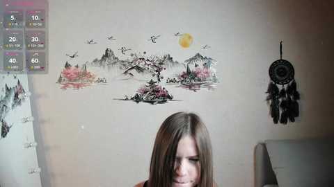 Video of a young woman with long brown hair, wearing a black top, looking down. Background features a wall mural with a mountain scene, a dreamcatcher, and a calendar.