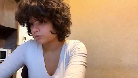 Video of a young woman with curly, shoulder-length black hair, wearing a white V-neck long-sleeve shirt, standing in a beige-walled kitchen with a microwave and stove in the background.