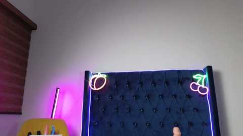 Video of a plush, dark blue, tufted headboard with neon green cherries and pink lighting on a light grey wall.