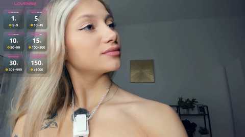 Media: Video of a young, light-skinned woman with long, straight blonde hair, wearing a silver necklace with a dog tag. She gazes upward, with a soft expression. Background shows a minimalist room with a plant and framed art.