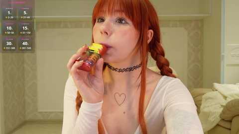 Video of a pale-skinned woman with red hair in braids, wearing a white top and black choker, eating a yellow candy in a beige room.