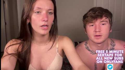 Video of a young woman with long brown hair, wearing a beige lace bra, and a man with short brown hair, shirtless with a laurel tattoo on his chest, both looking serious in an indoor setting.