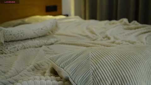 Video of a neatly made bed with cream-colored knitted blanket and pillow, beige walls, and dark curtains in the background.