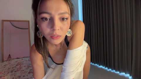Video of a young Asian woman with light skin, straight brown hair, wearing a white sleeveless top, leaning forward, and looking directly at the camera. She has large, round, blue earrings. Background shows a patterned bedspread, a wooden headboard, and vertical blinds.