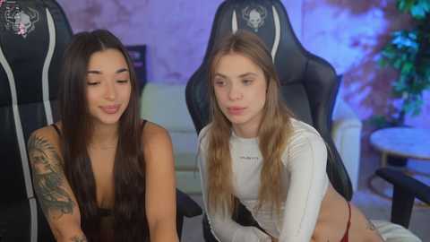 Media: Video of two young women with long hair, one with a sleeve tattoo, wearing low-cut tops, seated on gaming chairs in a cozy, dimly lit room.