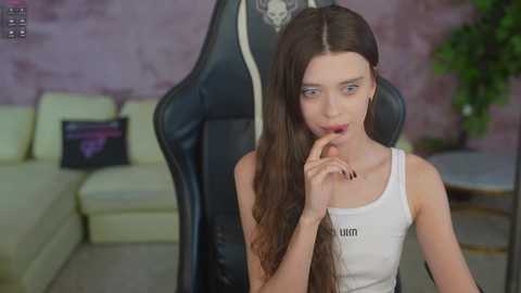 Media: Video of a slender, pale-skinned young woman with long brown hair, wearing a white tank top with \"Amanda\" printed, biting her index finger in a gaming chair against a purple wall.