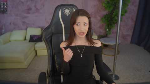 Video of a young woman with straight brown hair, wearing a black off-shoulder top, sitting on a gaming chair in a modern living room with a purple wall, green plant, and beige couch.