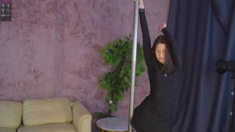 Video of an Asian woman with long brown hair in a black dress, performing a pole dance in a dimly lit room with a beige couch, textured pink wall, and green potted plant.
