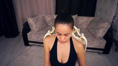 Video of a young woman with fair skin, dark hair pulled back, wearing a black halter top, sitting on a plush, beige couch in a dimly lit room.