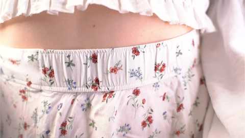 Media: A close-up video of a woman's midsection in a white floral dress with red, blue, and green flowers. The dress has a gathered elastic waistband and frilled neckline.
