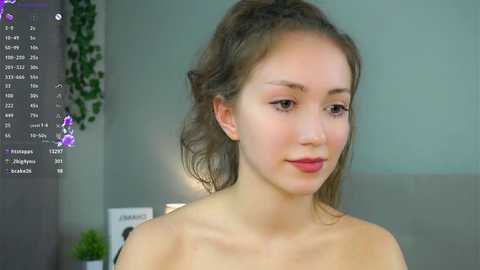 Video of a young, fair-skinned woman with light brown hair in a bun, wearing a strapless top, in a modern indoor setting with green plants.